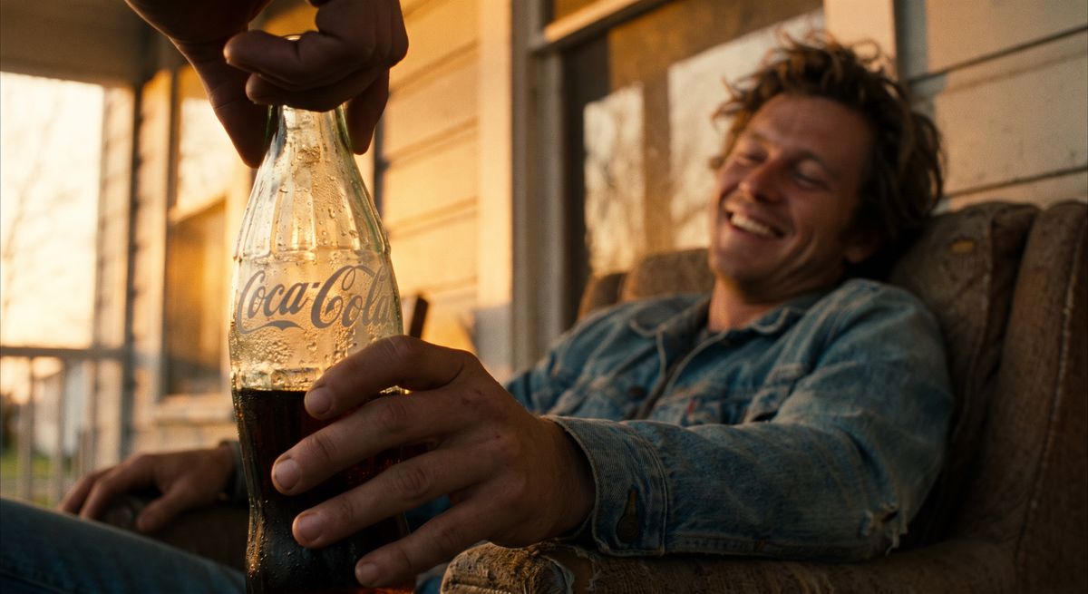 Panel 6 del storyboard. Mark leans back happily holding the condensation-covered glass bottle in the golden hour light, feeling fully at home.